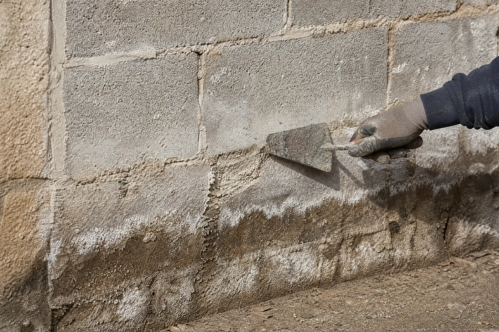 Truelle pointant le bas d'un mur en parpaing marqué d'un voile blanc et d'une bande sombre d'humidité.