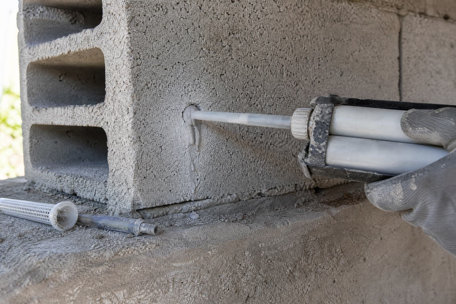 Pistolet à cartouche injectant de la résine dans un trou percé au bord d'un parpaing creux, mélangeur usagé posé devant.